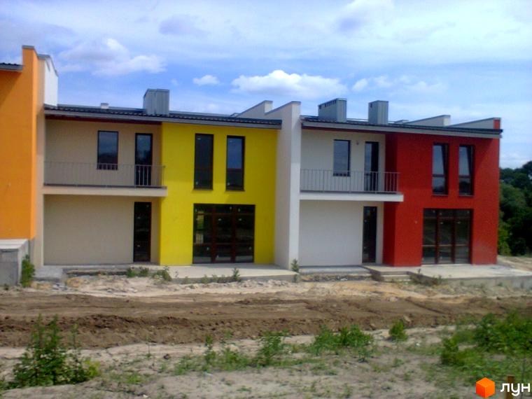 Ряд двоповерхових таунхаусів „Фільварок“ з різнокольоровими фасадами та темними вікнами. Перед будинками ґрунтова ділянка.