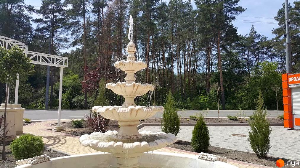 Житловий комплекс Карат. Фонтан у дворі з трьома ярусами, навколо якого викладена плитка. На задньому плані дерева та дорога.