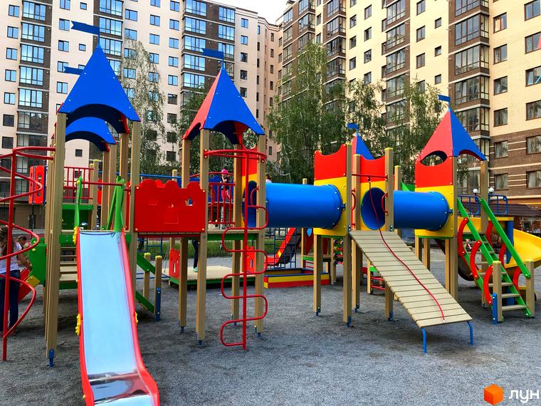 Фото дитячого майданчика у Житловому комплексі Андорра. На передньому плані різнокольорові гірки та ігрові конструкції, на фоні багатоповерхові будинки.
