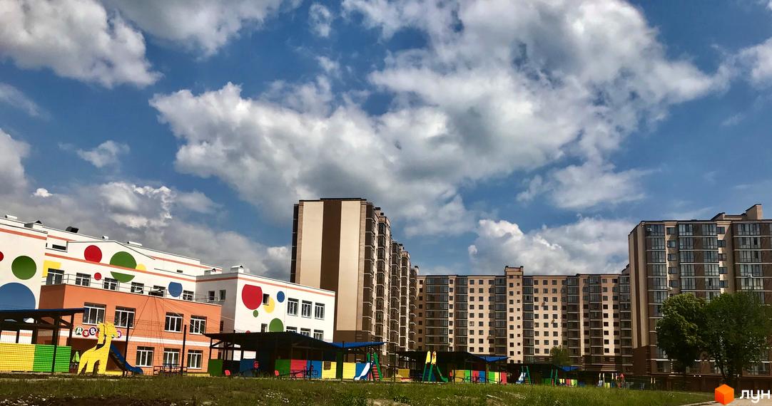 Зовнішній вигляд житлового комплексу Андорра з кольоровим дитячим майданчиком. На задньому плані висотні будинки та блакитне небо з хмарами.