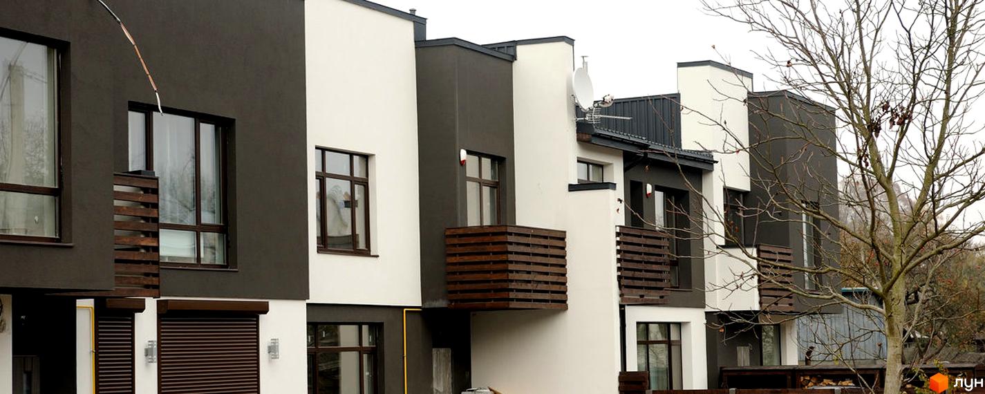 Зображення фасаду таунхаусів в ЖК «На Заозерній». Будинки з білими та темно-коричневими фасадами, дерев'яними балконами та великими вікнами. Поруч росте дерево.