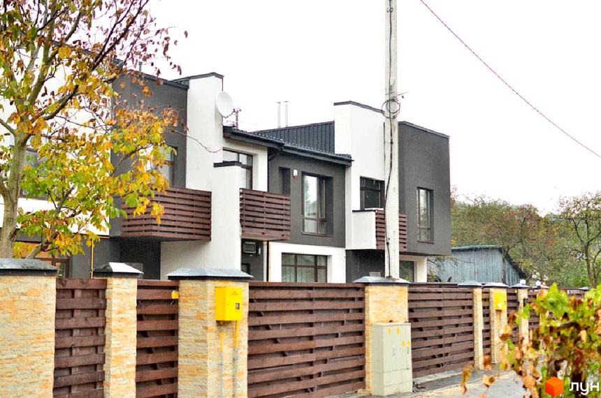 Зображення фасаду таунхаусів в житловому комплексі „На Заозерній“. Будинки з білими та сірими стінами, дерев'яними балконами та парканом з коричневих дощок.