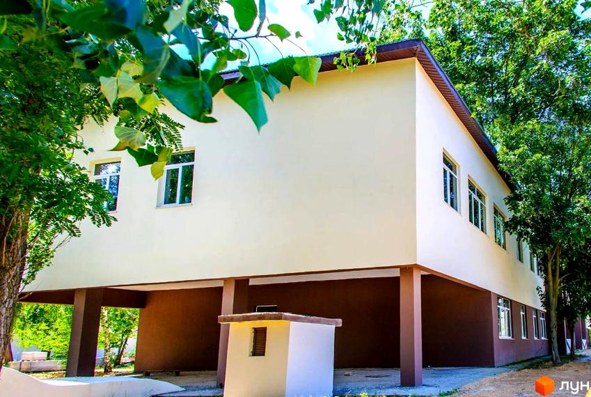 Новобудова The Loft з бежевими стінами на піднятому поверсі та коричневою основою, оточена зеленими деревами.