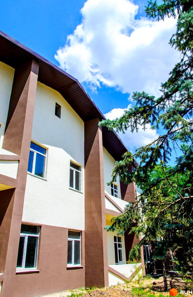 Зображення фасаду будинку житлового комплексу The Loft. Білі стіни з коричневими елементами, вікна з білими рамами. На тлі блакитне небо з хмарами та зелені дерева.