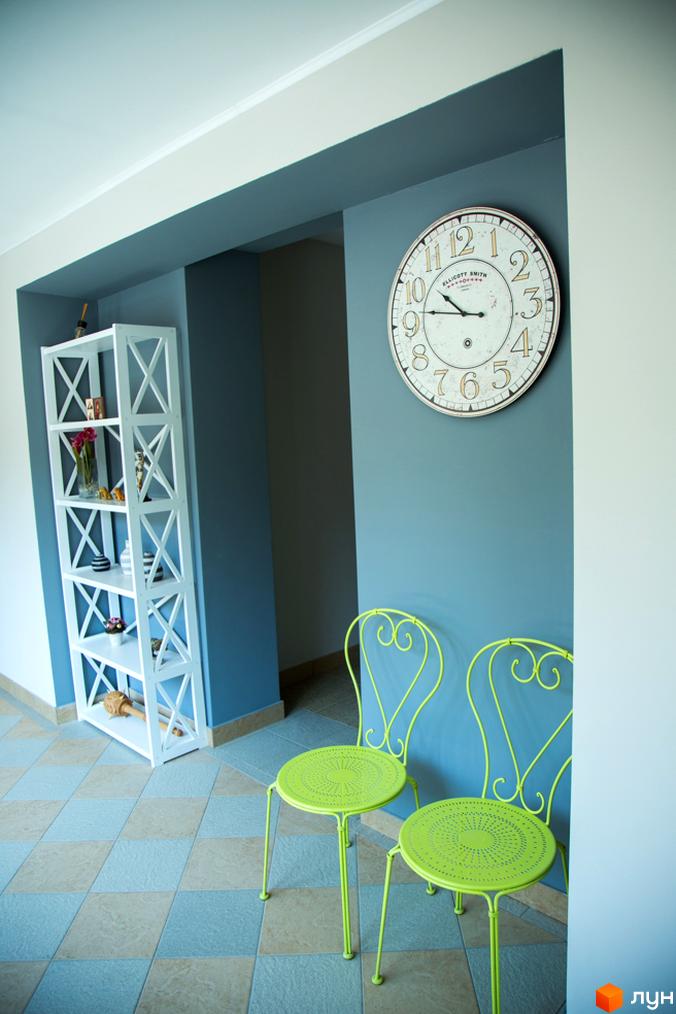 Інтер'єр житлового комплексу Comfort Club з білою стелажем, круглим годинником та двома зеленими стільцями. Плитка на підлозі у вигляді ромба.