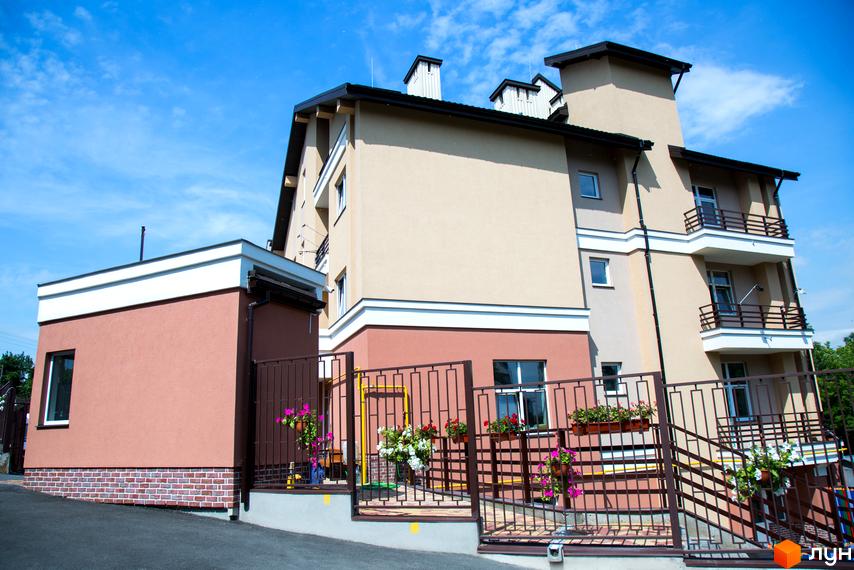 Житловий комплекс Comfort Club: фасади будівель в бежевих та рожевих тонах з білими елементами, балконами та коричневими огорожами. Прибудинкова територія з квітами у підвісних кашпо.