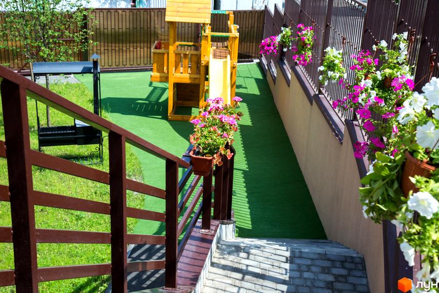 Житловий комплекс Comfort Club: дитячий майданчик з гіркою та квітами на прибудинковій території. Зелений газон та дерев'яні перила біля сходів.
