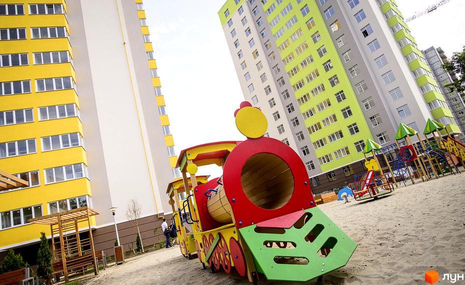 Житловий комплекс Відрадний: яскраві жовті та зелені фасади висотних будинків. На прибудинковій території дитячий майданчик у вигляді потяга та пісочниця.