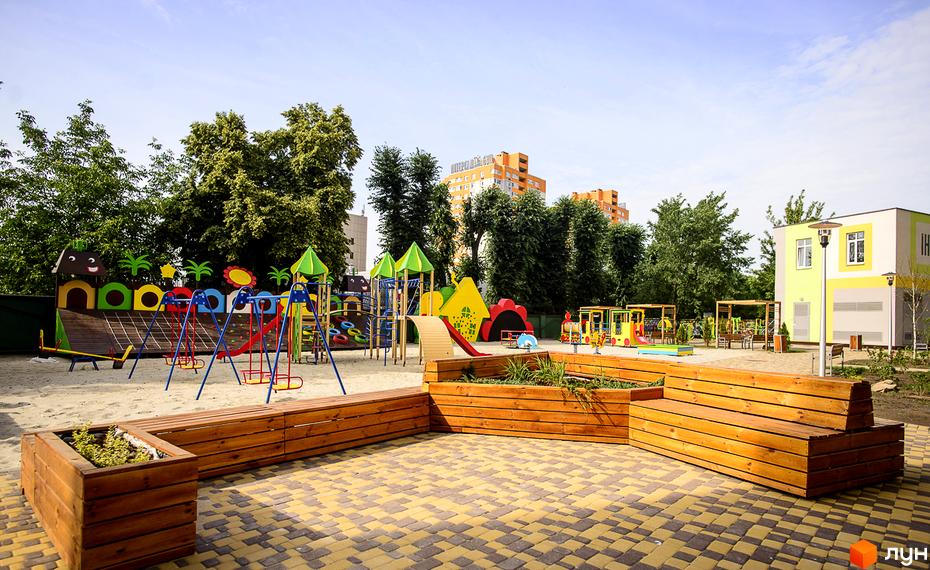 Будівельний проєкт Відрадний: дитячий майданчик з різнокольоровими гірками, гойдалками та пісочницею. На задньому плані – сучасні житлові будинки та зелені насадження.