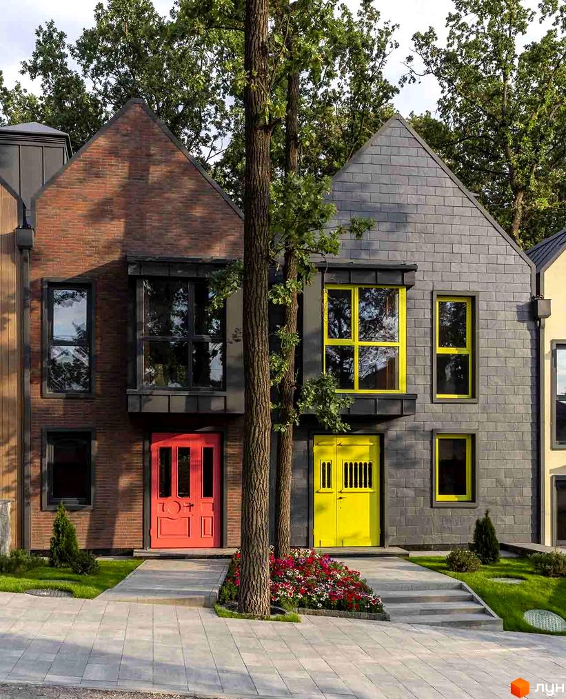 Зовнішній вигляд будинку житлового комплексу КМ Oak Grove Town з фасадом з цегли та сірого каменю. Перед будинком викладена плитка, ростуть квіти та дерева.