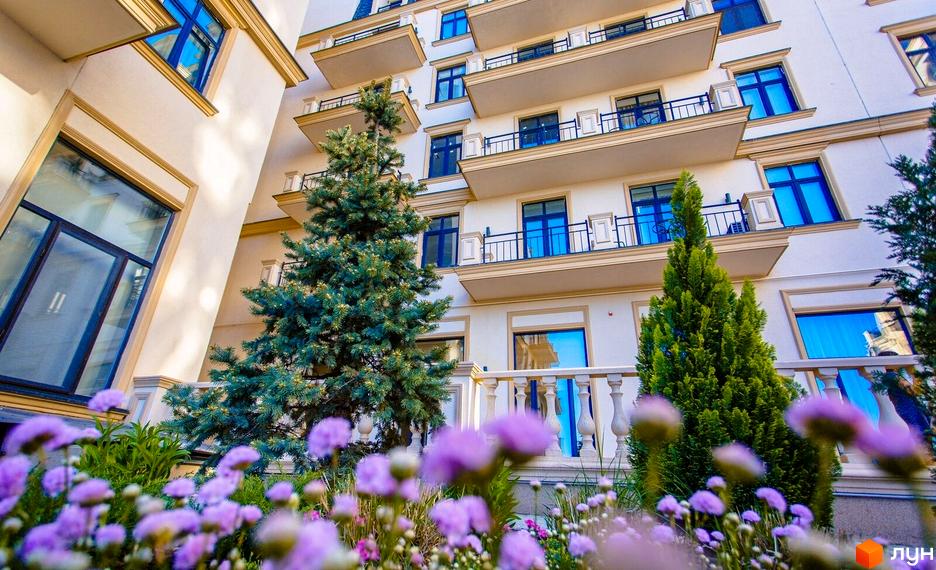 Житловий квартал Авторський: світлий багатоквартирний будинок з балконами та синіми вікнами, оточений зеленими деревами та фіолетовими квітами.