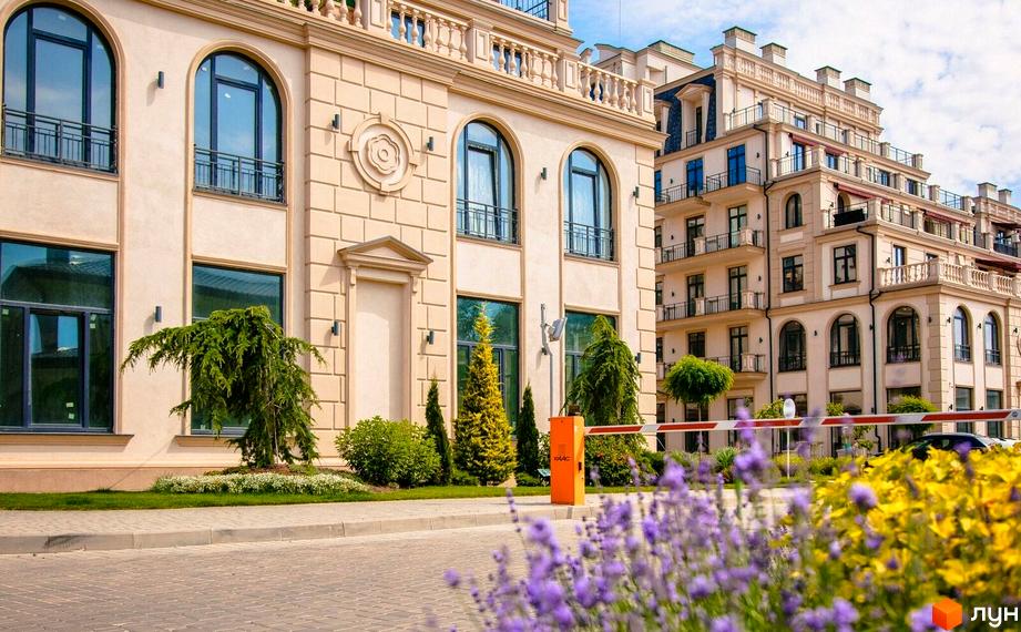 Фасад Житлового кварталу Авторський з бежевими стінами, арковими вікнами та класичними елементами. Прибудинкова територія з деревами, квітами та в'їзним шлагбаумом.