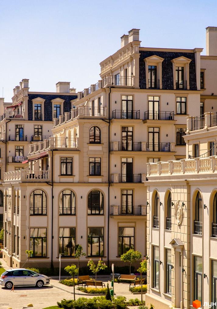 Світлі багатоповерхові будинки Житлового кварталу Авторський з темними мансардними дахами, балконами та арковими вікнами. Прибудинкова територія має зелені насадження, лавки та парковку.