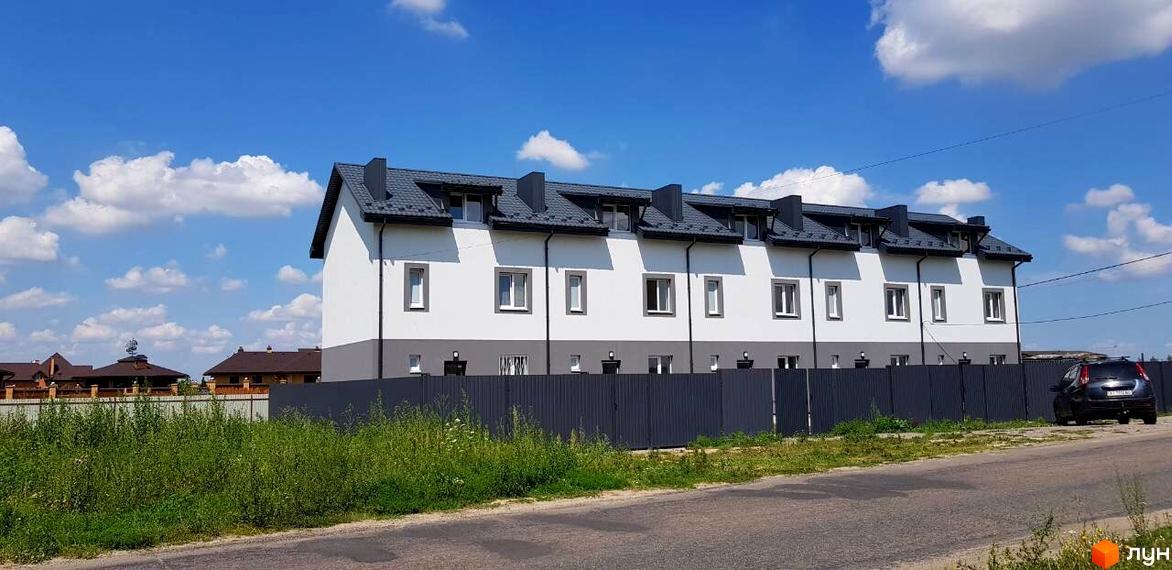 Таунхауси HarmonyBud: ряд білих таунхаусів з сірими дахами та вікнами, розташованих за темним парканом. Перед будинками зелена трава та дорога.