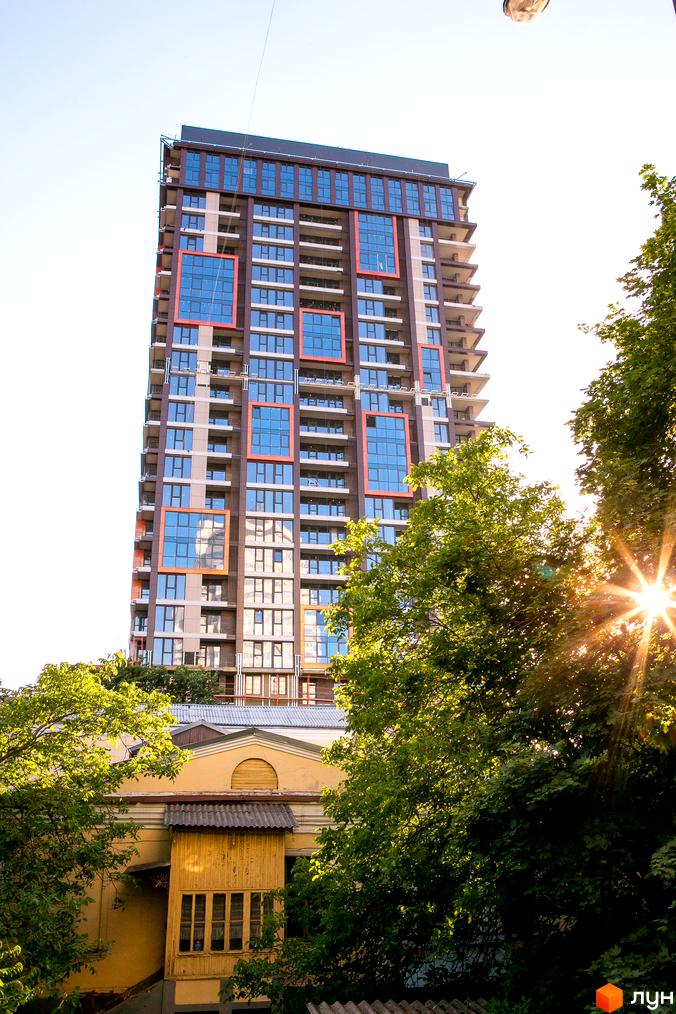 Вид на новобудову ЖК Новодворянський з фасадом з коричневих та сірих панелей та оранжевими квадратними вставками. Навколо зелені дерева та стара будівля.