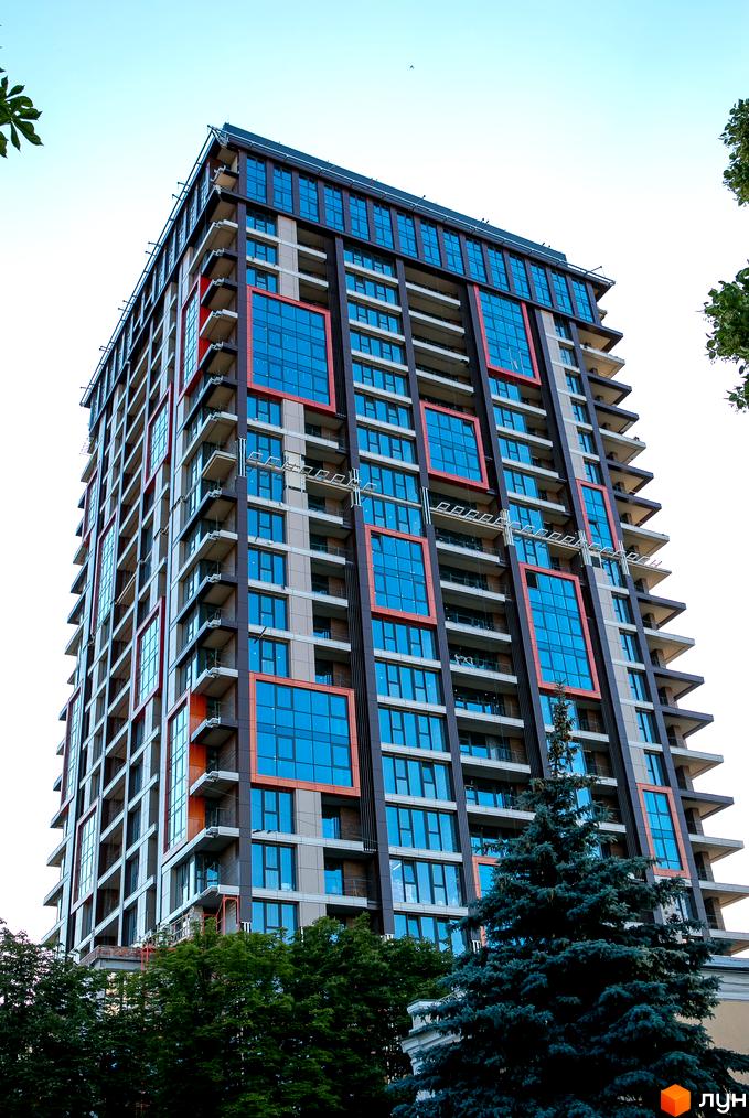 Зовнішній вигляд житлового комплексу Новодворянський з синіми вікнами та червоними елементами. Навколо будівлі зелені дерева та блакитне небо.