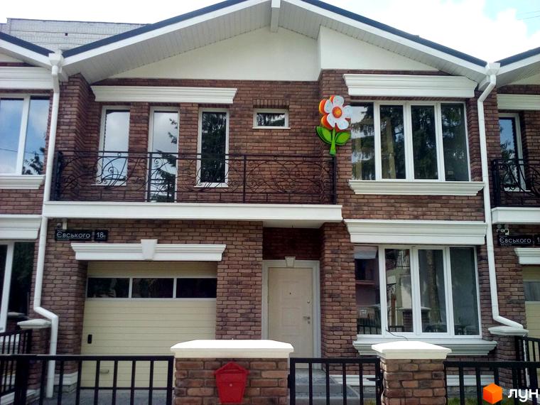 Фасад таунхауса на вул. Євського, 18 з коричневої цегли, білими вікнами, балконами та гаражем. Декоративна квітка на стіні, перед будинком чорна огорожа.