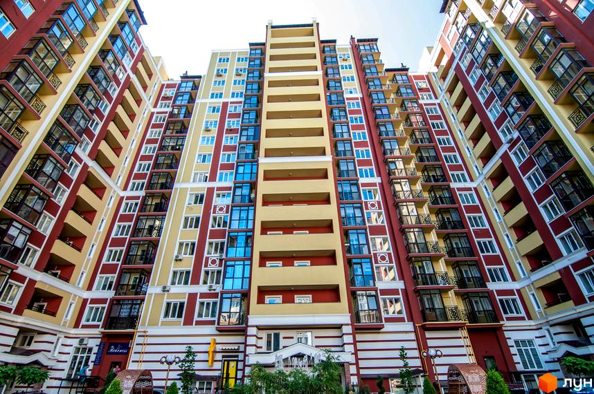 Зовнішній вигляд ЖК Версаль Парк: багатоповерховий будинок з червоно-жовтими фасадами та скляними балконами. Прибудинкова територія з зеленими насадженнями та елементами декору.