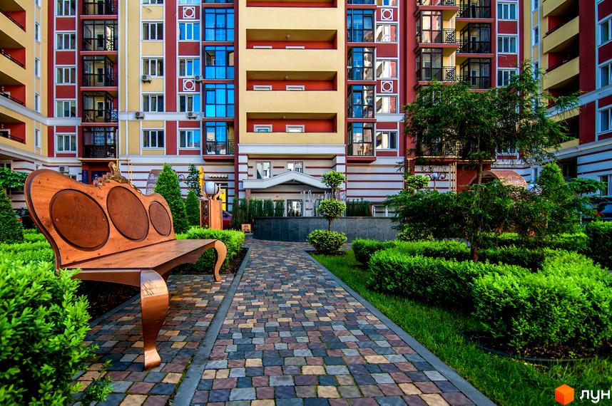 Житловий комплекс Версаль Парк: фасади будівель у червоних та жовтих тонах, упорядкована прибудинкова територія з доріжками, кущами та лавкою.