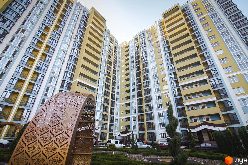 Житловий комплекс Версаль Парк - багатоповерхові будинки жовто-блакитного кольору з балконами та великою кількістю вікон. Прибудинкова територія з доглянутими кущами та декоративною аркою.