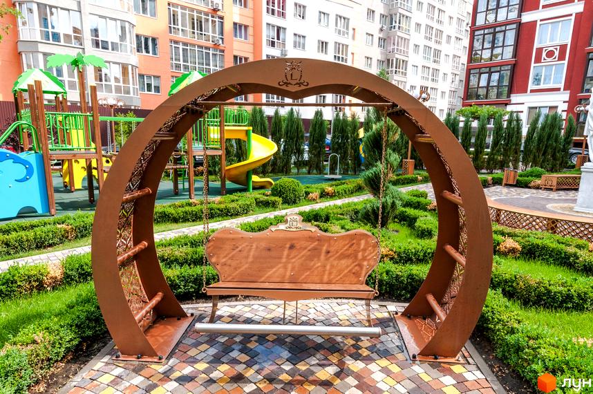 Житловий комплекс Версаль Парк: дитячий майданчик з гойдалкою та зеленими насадженнями. На задньому плані видно багатоповерхові будинки.