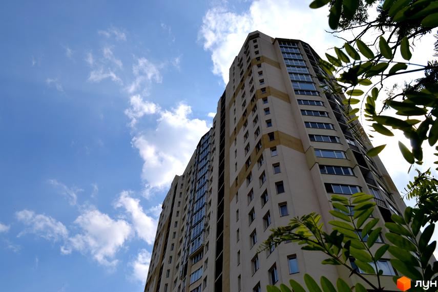 Висотний будинок житлового комплексу Ювілейний з великими вікнами на фоні блакитного неба. Прибудинкова територія з зеленими насадженнями.