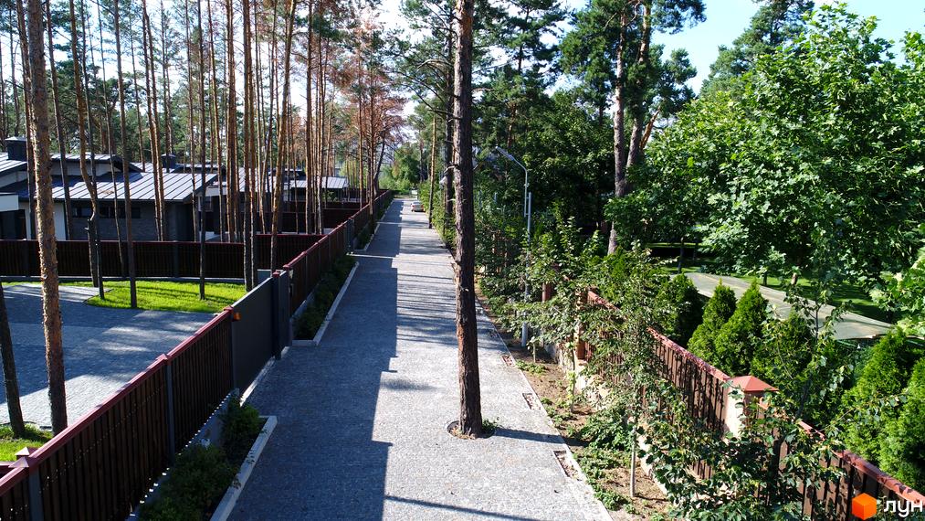 Дорога в Містечко котеджів Хвойний квартал. Цегляний паркан, дерева та котеджі з сірою покрівлею створюють атмосферу спокою та комфорту.