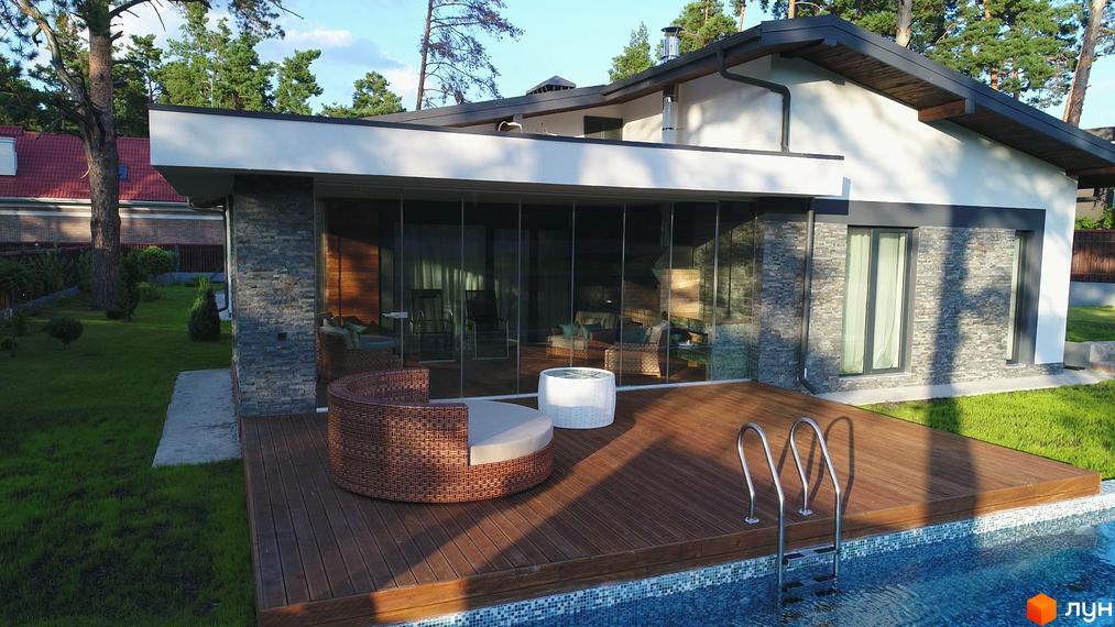 Зображення будинку з панорамними вікнами та терасою з басейном. На терасі розташовані меблі для відпочинку, навколо будинку зелена галявина та дерева.