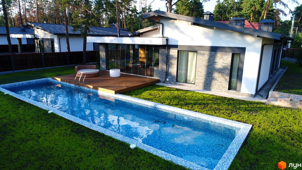 Котеджне містечко Хвойний квартал: білий будинок з кам'яною обробкою, великими вікнами. Поруч басейн, дерев'яна тераса та зелений газон.