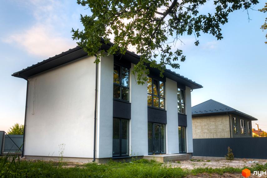 Зображення двоповерхового будинку в Котеджному містечку Lion`s Nest з великими вікнами та чорним дахом. Навколо будинку зелена трава та дерева.