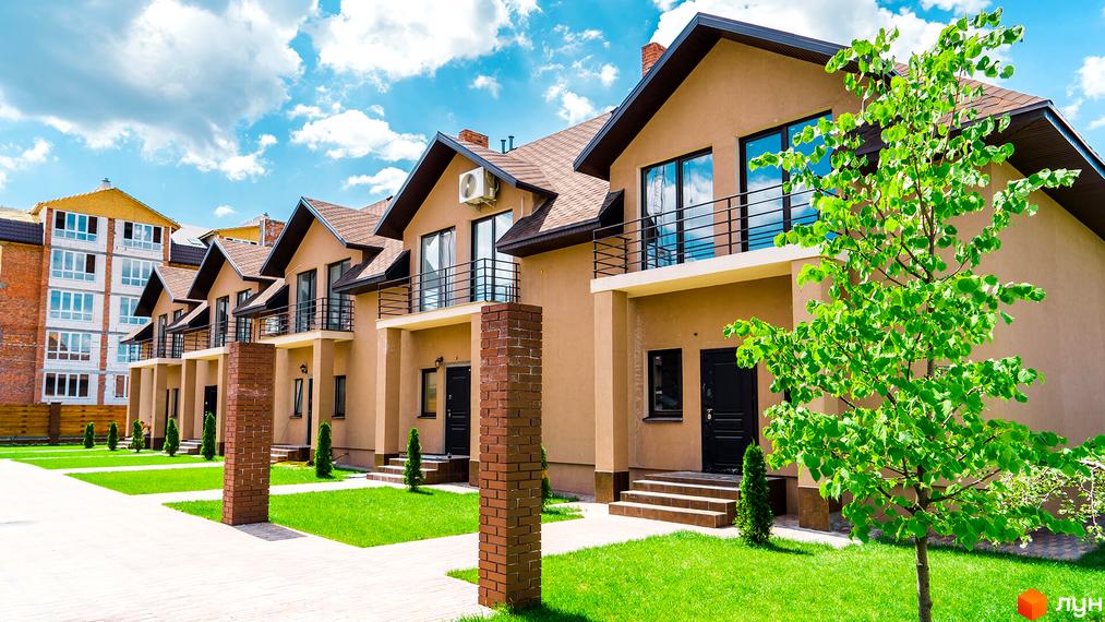 Ряд двоповерхових таунхаусів світло-коричневого кольору з темними дахами та балконами. Перед будинками зелені газони, доріжки та молоді дерева.