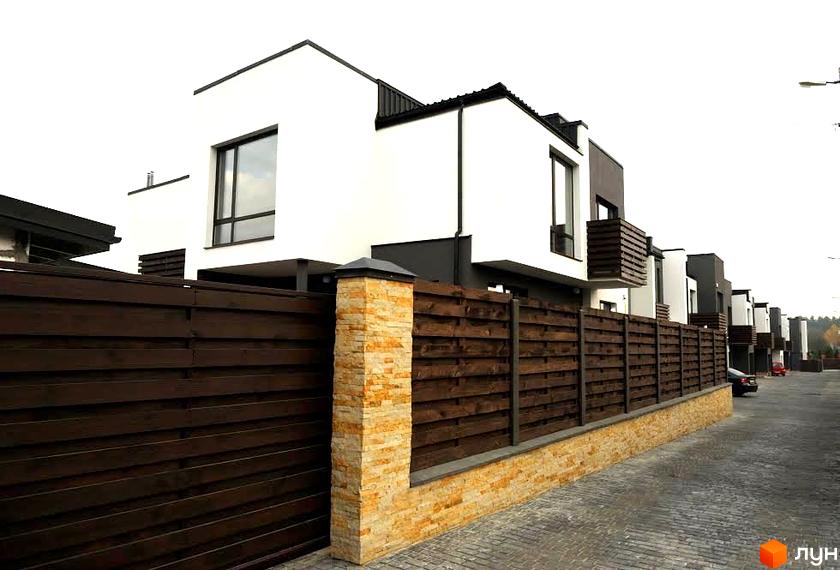 Зображення житлового комплексу. Білі будинки з темними дахами та вікнами, дерев'яний паркан з кам'яними стовпами. Бруківка на території комплексу.