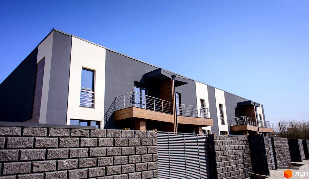 Зображення житлового комплексу Elegant. Будинки з балконами та фасадами у сіро-білих тонах. На передньому плані кам'яний паркан та металева огорожа.