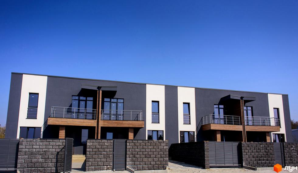 Зовнішній вигляд таунхаусів „Elegant“ з темними фасадами та балконами. Територія огороджена цегляним парканом з металевими воротами.