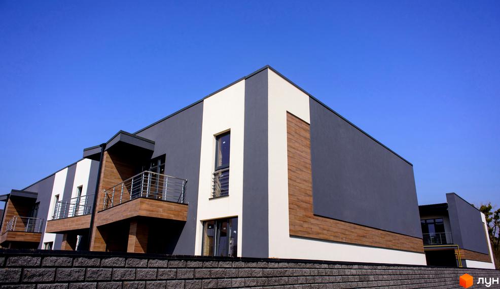 Зображення фасаду таунхаусів Elegant з темними стінами, дерев'яними вставками та балконами. Перед будинками розташований кам'яний паркан, а небо над ними - блакитне.