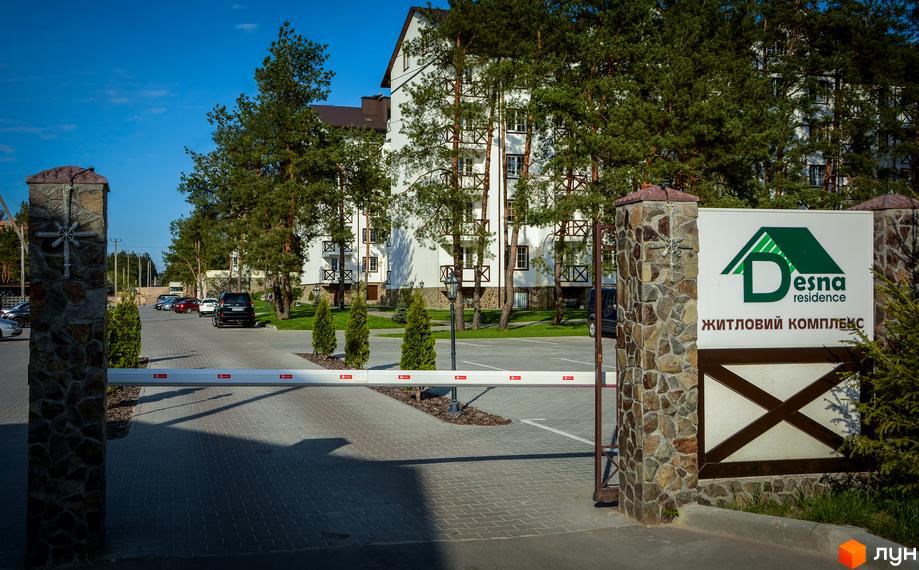 Житловий комплекс DESNA residence з білими фасадами та дерев'яними елементами, оточений соснами. Територія перед будинком облаштована парковкою та шлагбаумом.