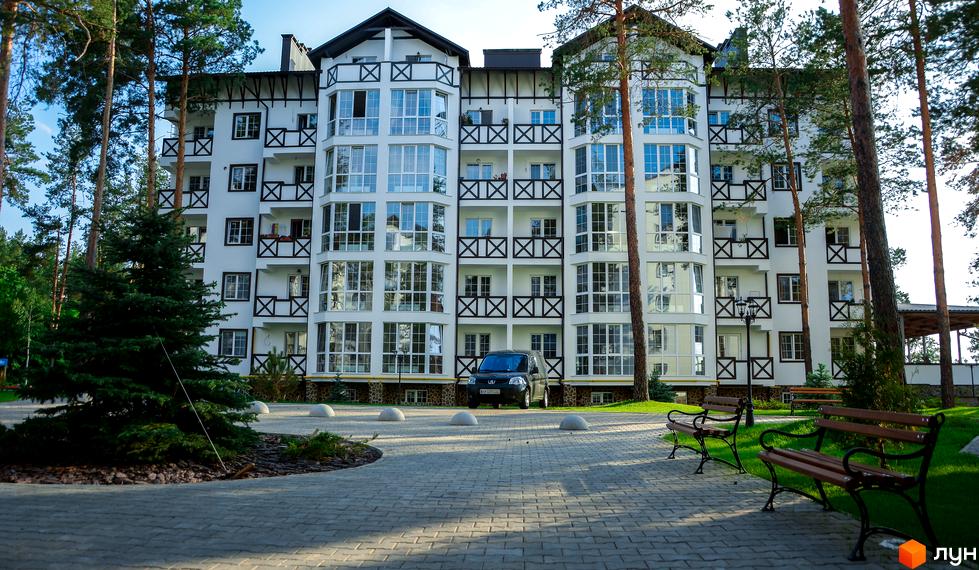 Житловий комплекс DESNA residence - біла багатоповерхівка з великою кількістю вікон та темними балконними елементами. Навколо облаштована територія з доріжками, газонами та деревами.