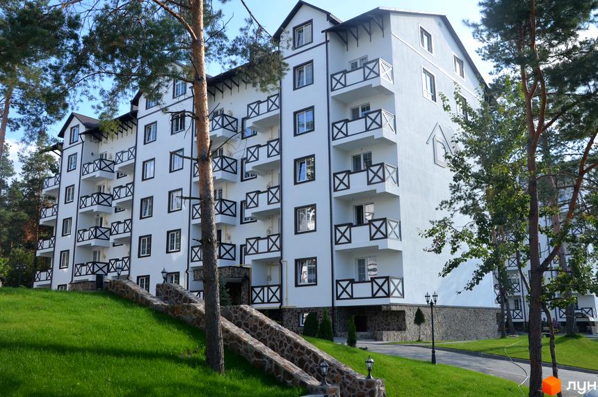 Житловий комплекс DESNA residence - біла багатоповерхова будівля з балконами та дерев'яними перилами, розташована на схилі серед дерев. Прибудинкова територія з доглянутим газоном та кам'яними сходами.