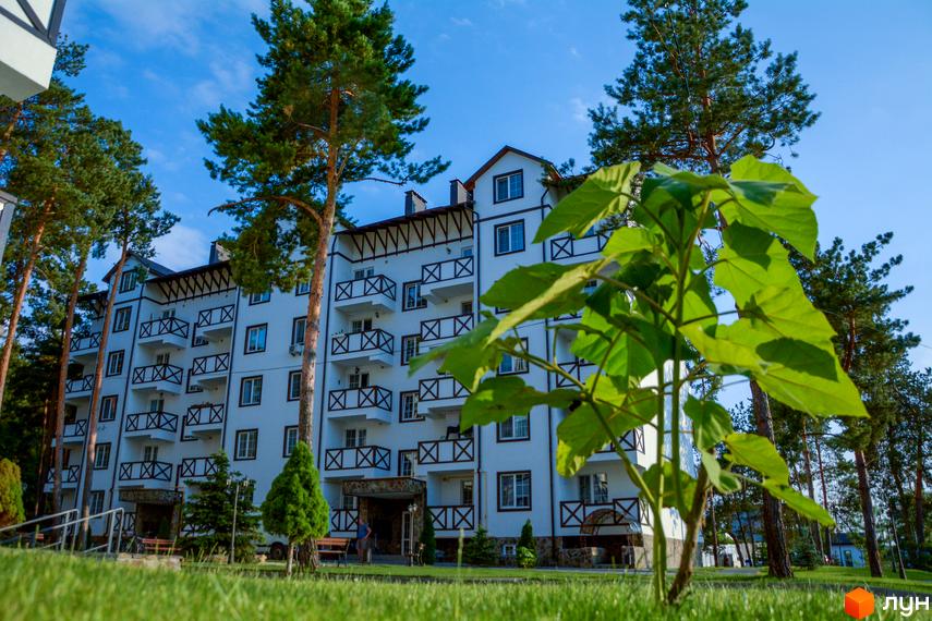 Житловий комплекс DESNA residence: біла багатоповерхова новобудова з балконами та соснами навколо. Зелена галявина та блакитне небо.