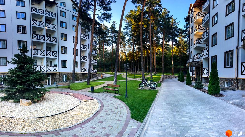 Житловий комплекс DESNA residence з білими фасадами та балконами з дерев'яними поручнями. Прибудинкова територія з доріжками з бруківки, зеленим газоном та деревами.