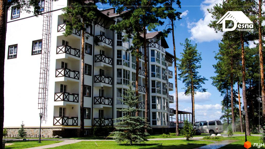 Житловий комплекс DESNA residence з білим фасадом, балконами з дерев'яними елементами та великими вікнами. Навколо комплексу зелена територія з деревами та доглянутим газоном.