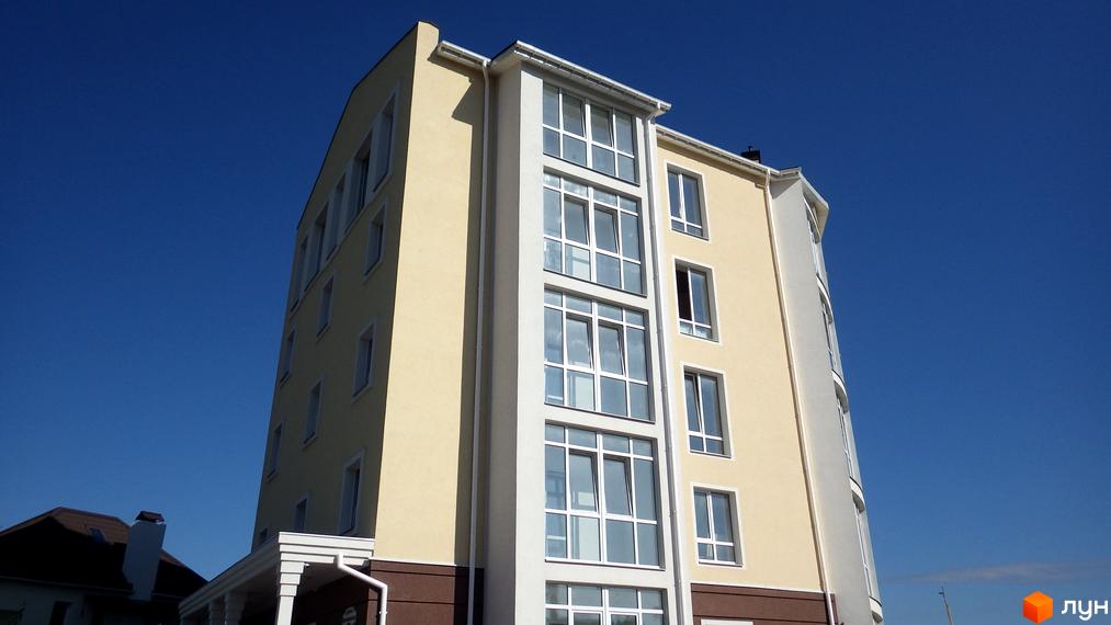 Висока будівля житлового комплексу Мандарин з великими вікнами та світлим фасадом на тлі блакитного неба. На території комплексу є зелені насадження.