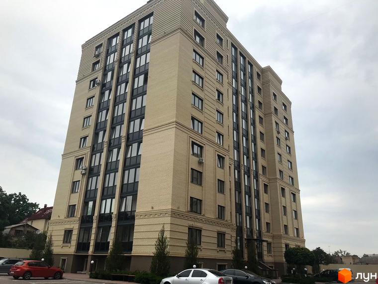 Висока новобудова з великою кількістю вікон. На прибудинковій території є парковка з автомобілями та зелені насадження.