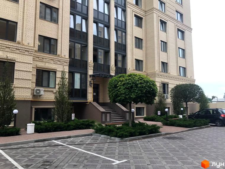 Зовнішній вигляд будівлі ЖК Центральний з світлої цегли з великими вікнами. На прибудинковій території висаджені кущі та дерева, є парковка.