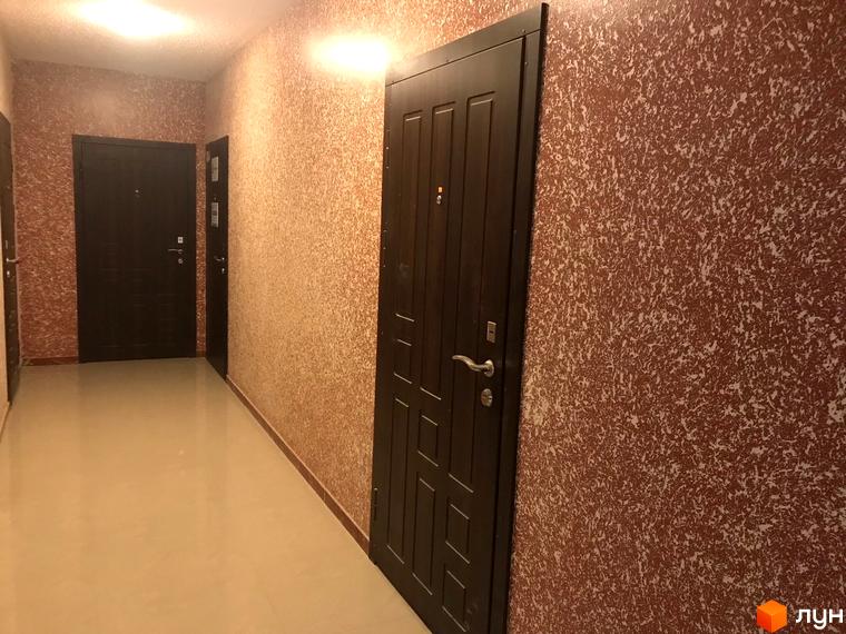 Коридор житлового комплексу з дверима темного кольору та стінами з декоративним покриттям. На підлозі світла плитка.