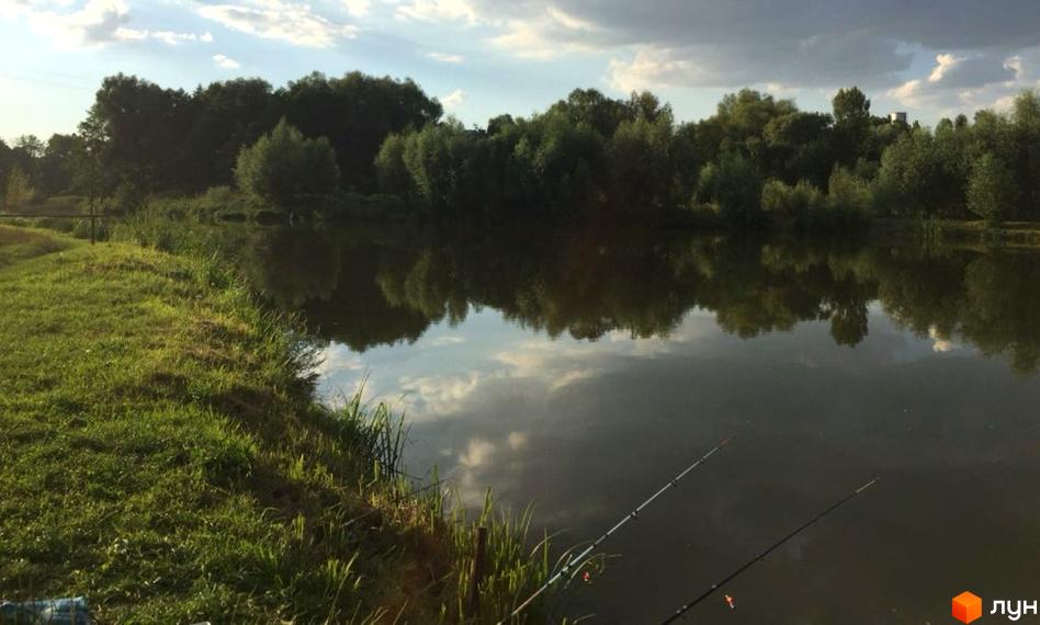 Котеджний квартал „Гетьманський 3“ – мальовничий вид на річку з берегом, вкритою травою, та деревами на протилежному березі. На передньому плані видно вудки для риболовлі.