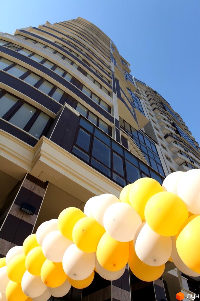 Висотна будівля Житлового комплексу Престиж Холл з фасадами темно-коричневого та світлого кольорів, та численними вікнами. На передньому плані арка з жовтих та білих повітряних кульок.