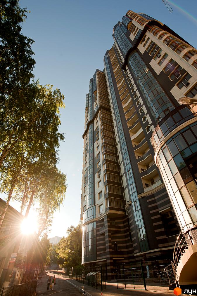 Багатоповерховий Житловий комплекс Престиж Холл зі скляними фасадами та балконами, розташований поруч із зеленими деревами та пішохідною вулицею під сонячним небом.