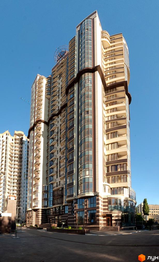 Житловий комплекс Престиж Холл: висотна будівля з бежевими та коричневими фасадами, великими вікнами та балконами. Перед будинком дорога та зелені насадження.