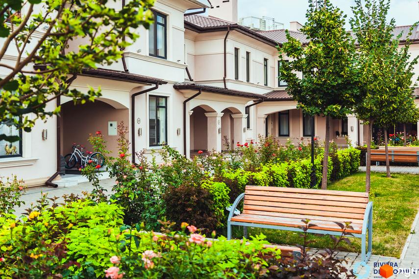 Таунхауси „Вілла Троянд“ з рожевим фасадом, великими вікнами та коричневим дахом. Навколо будинку зелені насадження, квіти та лавки для відпочинку.